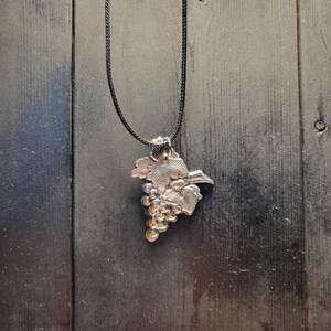 May include: Silver-toned pendant necklace featuring a detailed grape cluster design with leaves. The pendant hangs from a black cord. The necklace is displayed against a dark wooden background.