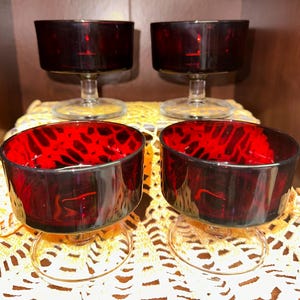 Op de afbeelding: Vier vintage rode glazen dessertkommen met heldere stelen. De kommen hebben een donkerrode buitenkant en een lichtere, gespikkelde rode binnenkant. De kommen zijn gerangschikt op een gele kanten placemat, met een donkerbruine achtergrond.