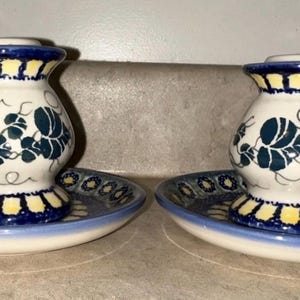 May include: Two matching ceramic candle holders with handles, each set on a matching saucer. The candle holders are white with blue and yellow accents and a blue floral design. The saucers are blue and white.