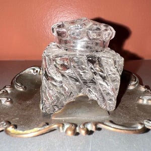 Puede incluir: Un tintero de cristal transparente con un diseño en espiral se encuentra sobre una bandeja decorativa plateada. La bandeja tiene una forma adornada y curvada con un borde de color bronce. El tintero tiene una tapa a juego.