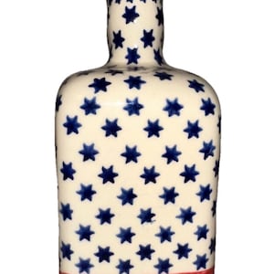 May include: A ceramic bottle with a cork stopper, featuring a white background adorned with blue stars and red and white stripes at the base. The bottle has a rounded rectangular shape.
