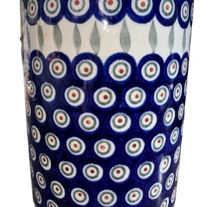 May include: A cylindrical ceramic mug featuring a repeating pattern of dark blue circles with white and light green centers, accented by small orange dots. The upper rim displays a decorative border of blue circles and green leaf-like shapes.
