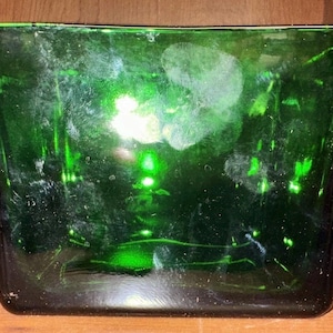 May include: A rectangular, dark green glass container with rounded corners. The glass has a glossy finish and appears to be slightly translucent, with some imperfections visible on the surface. The container is empty and sits on a wooden surface.