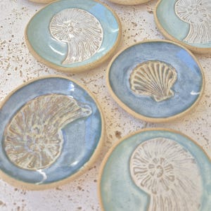 May include: A collection of seven small, round ceramic dishes with irregular edges. Each dish features a unique, embossed seashell or ammonite fossil design in white against a glossy blue or light green glazed background with tan rims.