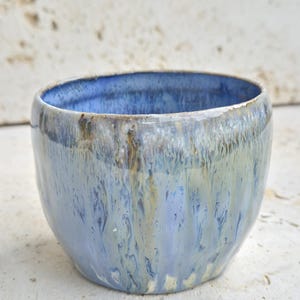 Könnte beinhalten: Eine kleine, runde Keramikschale mit einer glänzenden Glasur in einer Mischung aus Hellblau, Creme und Dunkelbraun. Die Glasur weist ein vertikales Tropfmuster auf, das die Außenfläche hinunterläuft, während das Innere einfarbig blau ist.