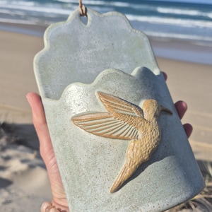 May include: A wall-mounted ceramic pocket planter with a light blue-green speckled glaze. The front features a raised, textured relief of a hummingbird in a tan color. A small hole at the top allows for hanging with an attached thin leather cord.