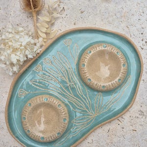 May include: An irregular, light blue ceramic tray featuring two circular, beige-coloured indentations with dotted rims. The surface is decorated with etched line drawings of wildflowers and stems, with the natural clay edges left exposed.