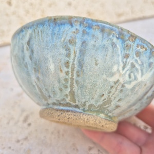 Pode incluir: Uma tigela de cerâmica feita à mão com um esmalte azul claro e branco. A tigela tem uma forma arredondada e uma superfície texturizada. A base é sem esmalte e tem um tom natural e terroso. Esta tigela única é perfeita para servir comida ou como peça decorativa.