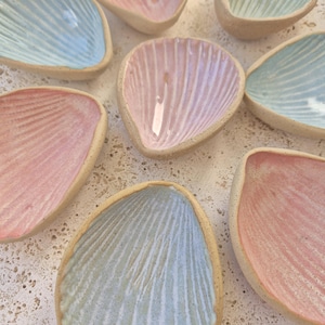 May include: A collection of eight small, shell-shaped ceramic dishes with a textured, unglazed exterior rim. The interior surfaces are finished in a glossy glaze featuring pastel shades of light blue, soft pink, and lavender with ribbed details.
