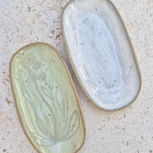 May include: Two oval ceramic trays with a speckled finish. One tray is light green and the other is off-white. Both feature an embossed botanical design of stems and flowers on the interior surface with exposed clay edges and a smooth glaze.