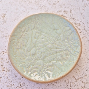 May include: A round ceramic dish with a light green glaze and a textured surface featuring embossed tropical leaf patterns. The edges of the dish are slightly raised, and the base material appears to be a natural, unglazed tan clay.