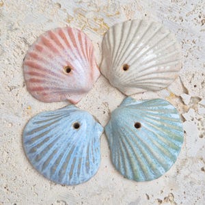 May include: Four ceramic seashell ornaments arranged in a square formation. Each shell features a glossy finish, radial ridges, and a single small circular hole near the top. The colors include a soft pink, a creamy off-white, a light blue, and a seafoam green.