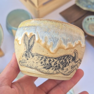Może przedstawiać: Mała ceramiczna miseczka z nakrapianą beżową podstawą i czarnym rysunkiem liniowym biegnącego królika. Górna krawędź posiada dekoracyjne szkliwo ściekające w odcieniach bieli, jasnego błękitu i żółci, co nadaje zewnętrznej stronie teksturę.