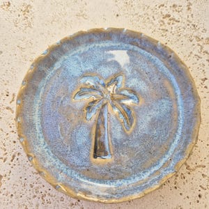 May include: A round ceramic dish featuring a raised palm tree design in the centre. The surface has a mottled glaze with shades of blue, grey, and tan, with a scalloped, textured edge that transitions into a warm, earthy brown tone.