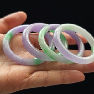 May include: Four jade bangles in shades of green and purple. The bangles are stacked on top of each other and are smooth and polished.