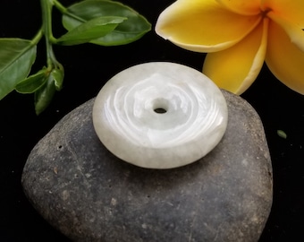 May include: A white jade pendant with a circular shape and a hole in the center. The pendant is resting on a gray rock.