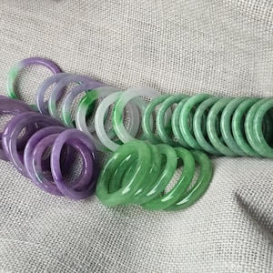 May include: A collection of jade bangles in shades of green and purple. The bangles are stacked and arranged on a textured fabric surface.