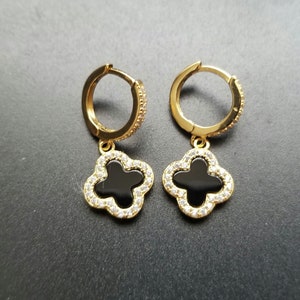 Peut inclure: Une paire de boucles d'oreilles créoles dorées avec un pendentif trèfle noir. Le pendentif trèfle est orné de pierres de zircone cubique transparentes.