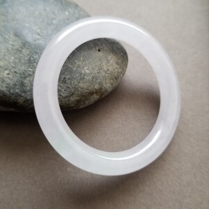 Puede incluir: Un brazalete de jade blanco translúcido. El brazalete circular se muestra sobre una piedra gris y un fondo neutro. El jade tiene una sutil variación de color natural. El diámetro del brazalete es de aproximadamente 6,5 cm.