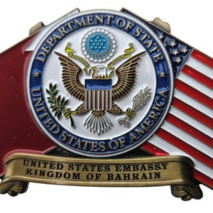 May include: A gold-colored metal pin with the United States Department of State seal, featuring a bald eagle with outstretched wings, surrounded by a blue circle with the words "Department of State" and "United States of America". The seal is flanked by a red and white American flag and a red flag. Below the seal is a gold banner with the text "United States Embassy Kingdom of Bahrain".
