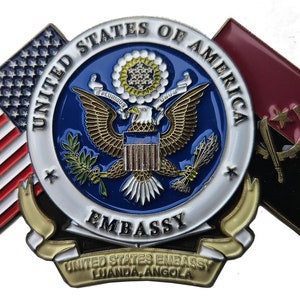 May include: A gold-colored pin with the United States seal and the words "United States of America Embassy" and "Luanda, Angola". The seal is surrounded by an American flag on the left and an Angolan flag on the right.
