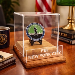 May include: A commemorative coin displayed in a clear acrylic case. The coin features the FBI seal and the words "New York Field Office." The display sits on a wooden base with "FBI NEW YORK CITY" etched on the front. A mug with the FBI logo is visible.