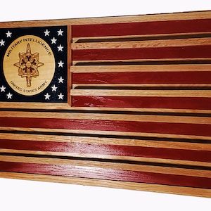 May include: A wooden American flag display with a red, white, and blue design. The flag has a circular emblem in the upper left corner that reads "Military Intelligence United States Army". The flag has eight slots for displaying medals or other items.