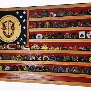 May include: A wooden shadow box with a red, white, and blue American flag design. The shadow box has six shelves for displaying challenge coins. The top shelf has a round emblem with the text "5TH SPECIAL FORCES GROUP DE OPPRESSO LIBER UNITED STATES ARMY".
