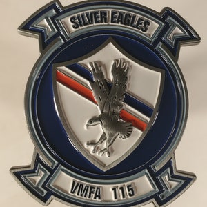 May include: A silver metal coin with a blue and white shield featuring a silver eagle. The shield is surrounded by a blue circle with the words "SILVER EAGLES" in a silver arc above the shield and "VMFA 115" in a silver arc below the shield.
