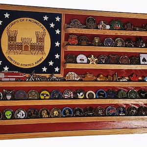 May include: A wooden shadow box with a red, white, and blue American flag design. The box has six shelves for displaying challenge coins. The top shelf features a round emblem with the words "Corps of Engineers, United States Army".