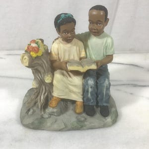 May include: A figurine of two children sitting on a tree stump reading an open book. The girl wears a cream dress with a teal headband, and the boy wears a light green shirt and blue jeans. A cluster of small orange, yellow, and white flowers sits atop the stump.