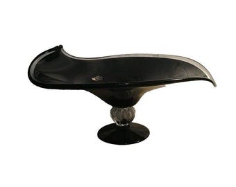 May include: A black glass bowl with an elongated, wave-like rim and a clear, ribbed glass stem. The bowl features a smooth, dark surface and sits on a circular black base. A small, rectangular label is visible on the interior of the bowl.