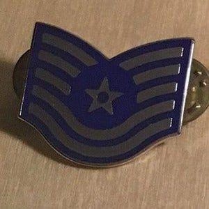 May include: A blue and gray enamel pin featuring a star and stripes design. The pin has a military theme and is likely a rank insignia. The pin is made of metal and has a pin back.