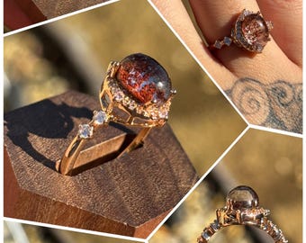 May include: A rose gold ring featuring a central oval-shaped phantom quartz with reddish-brown inclusions, surrounded by a halo of small clear stones. The band is accented with additional clear stones and labelled 44474 Phantom Quartz.