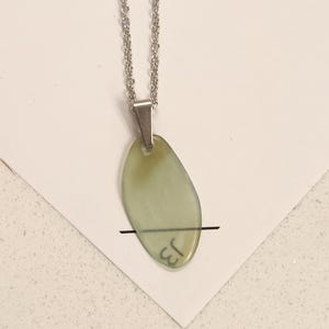 May include: A silver-toned necklace with a pendant. The pendant is an oval shape with a pale green hue. The pendant has the handwritten letters "J3" on it. The necklace is displayed on a white surface.