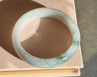 May include: A jade bangle bracelet. The bracelet is a pale green color with areas of darker green marbling. The smooth, polished surface reflects light, highlighting the natural variations in the stone. The bangle is resting on a light pink surface.