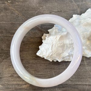 May include: A smooth, polished, translucent lavender-toned bangle bracelet with a rounded profile. The circular band features subtle variations in color, ranging from pale white to soft purple, and has a glossy, reflective surface finish.