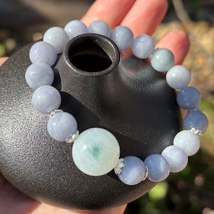 Puede incluir: Un brazalete de cuentas de jade azul pálido y verde. El brazalete tiene una cuenta central grande y cuentas más pequeñas con detalles plateados. El brazalete se exhibe sobre un jarrón de cerámica negro.