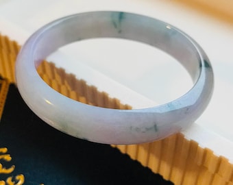 May include: A pale lavender jade bangle with subtle green and white marbling. The bangle is smooth and polished, and it is displayed in a white box.