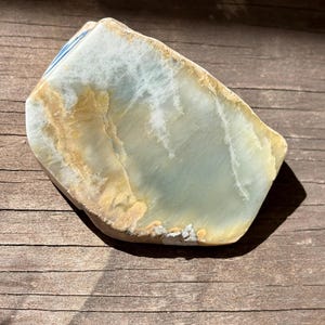 May include: A polished, irregular stone fragment featuring a smooth, translucent surface with swirling bands of pale blue, cream, and golden-brown hues. The edges are rounded and weathered, showing a natural, layered texture with a small blue and white ceramic shard visible at the top corner.