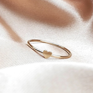 May include: A delicate gold ring with a small heart-shaped design. The ring is on a soft, beige fabric background.