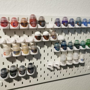 Peut inclure: Un panneau perforé blanc avec différentes petites bouteilles de peinture de couleurs variées, organisées en rangées. Les bouteilles sont étiquetées "Citadel Colour" et sont maintenues en place par des supports en plastique blanc.