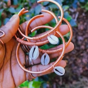 May include: Copper-colored metal bracelets with a spiral wire design and white seashell accents. The bracelets are open-ended and held in a hand, showcasing their handmade quality. The jewelry is set against a blurred green background.