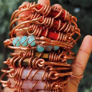 May include: A stack of copper wire bracelets featuring various gemstones. The bracelets include red rectangular stones, light blue round beads, brown striped stones, and pale pink rectangular stones, all secured with intricate copper wire wrapping.