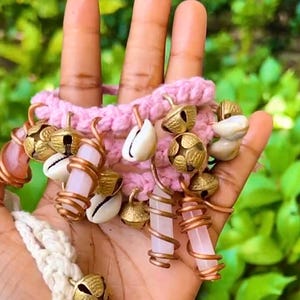 May include: A pink crocheted bracelet adorned with small gold bells, white seashells, and rose quartz crystals. The bracelet is accented with copper wire and a cream-colored macrame cord, creating a bohemian aesthetic.