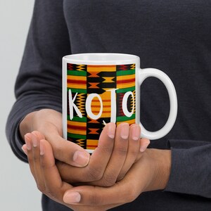 May include: White ceramic mug with a black, yellow, green, and red patterned design. The word "KOJO" is printed in white on the mug.