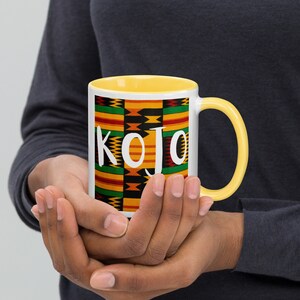 Pode incluir: Uma caneca de cerâmica branca com uma borda e uma alça amarelas. A caneca apresenta um design de estampa africana colorido com o nome "KOJO" em letras brancas.