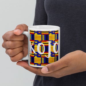 Könnte beinhalten: Weiße Keramiktasse mit einem farbenfrohen afrikanischen Druckmuster und dem Namen "KOJO" in weißen Buchstaben.
