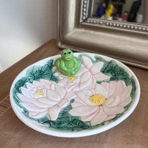 以下が含まれることがあります： ピンクと白の蓮の花と緑の葉のデザインの上に、緑のカエルの置物があるセラミック装飾皿。 皿は白い縁があり、木製の表面に置かれています。