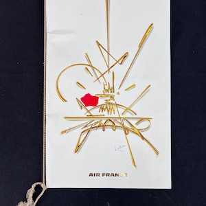 Könnte beinhalten: Eine Vintage-Broschüre von Air France mit einem weißen Umschlag, der ein goldenes abstraktes Design und einen roten Akzent aufweist. Die Broschüre ist mit einer beigen Schnur gebunden, darunter steht "AIR FRANCE".